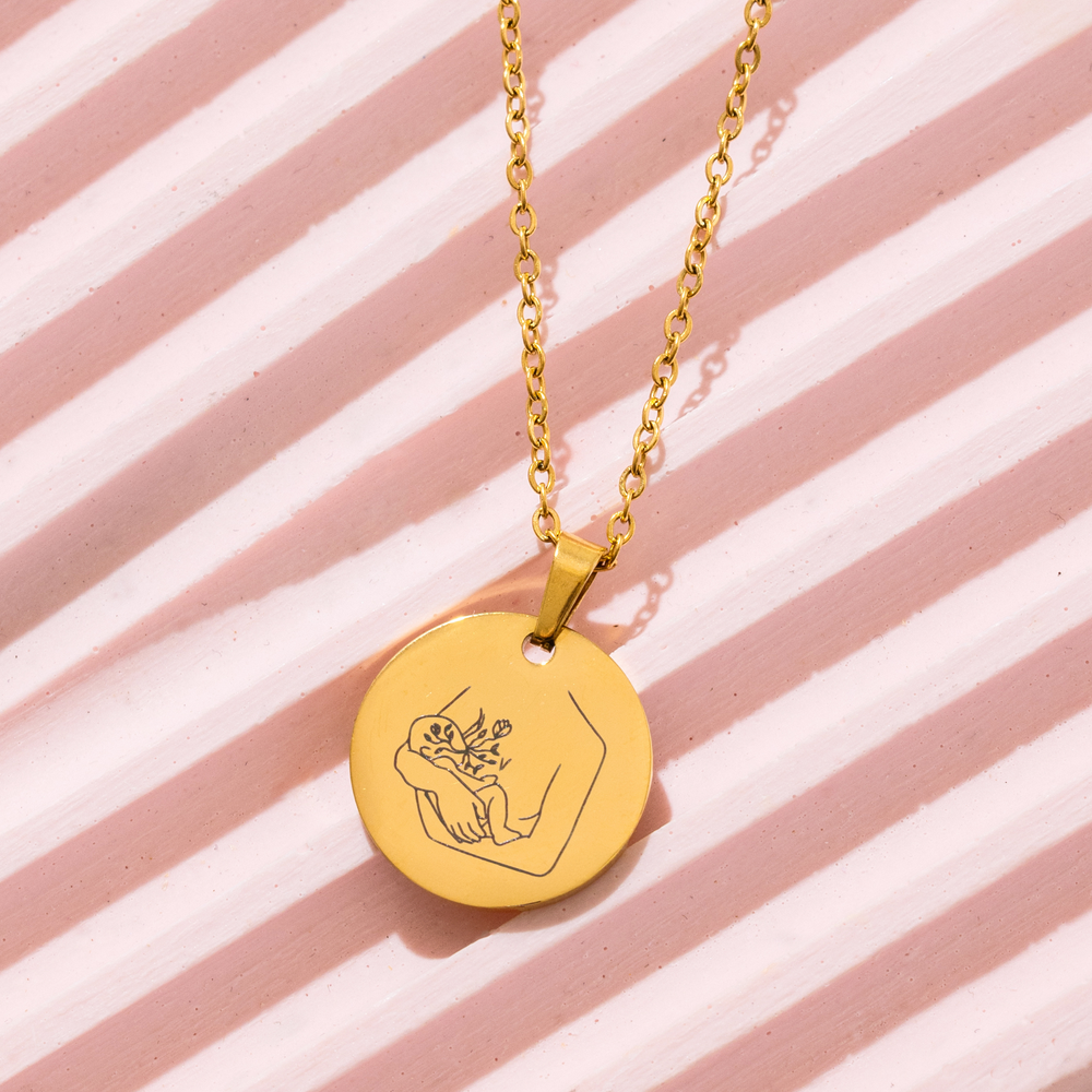 [main] A gold  pendant necklace with a gold chain featuring a mother breastfeeding her child. 